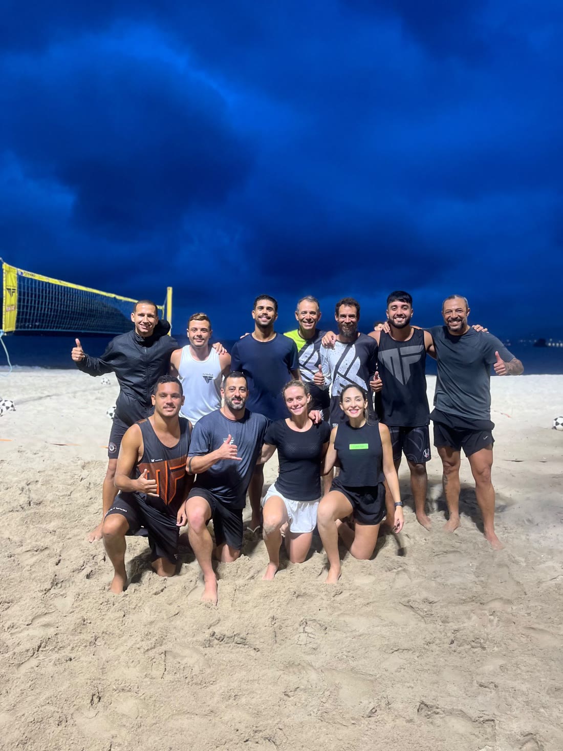 Professores e alunos da MF Futevôlei reunidos na quadra de areia ao anoitecer
