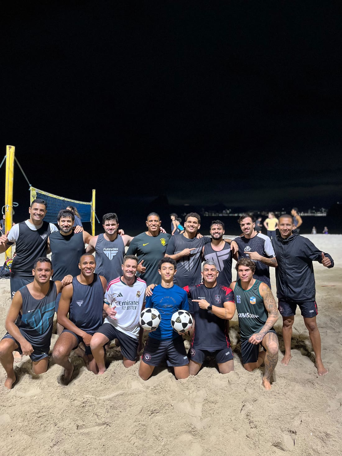 Turma da MF Futevôlei segurando duas bolas na quadra iluminada à noite